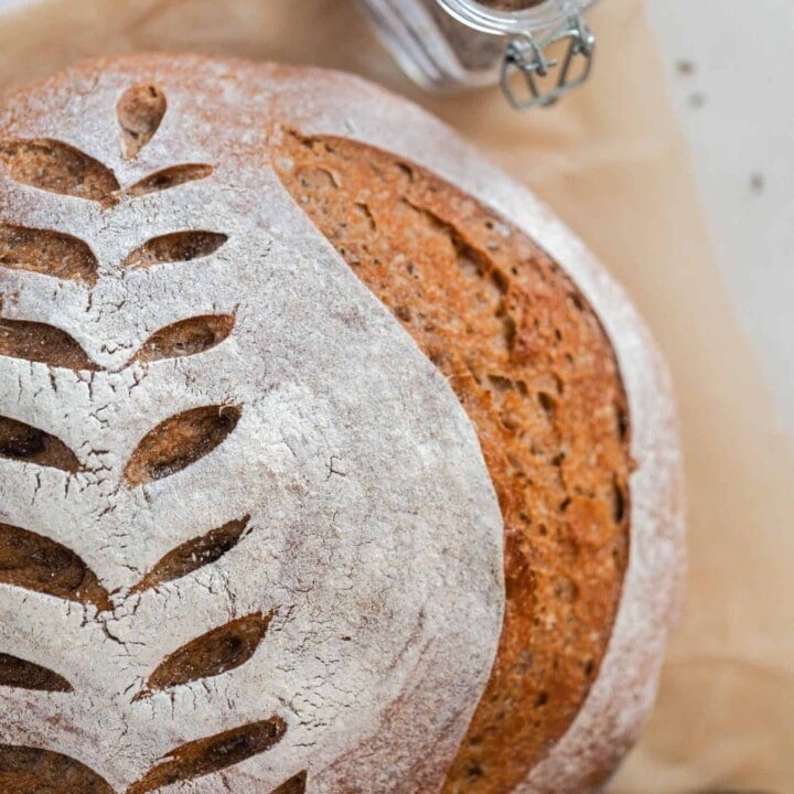 overhead photo of a crusty loaf of sourdough rye bread with a wheat design scored on top. The loaf rest on parchment paper with a jar of spices and a jar of molasses around the loaf