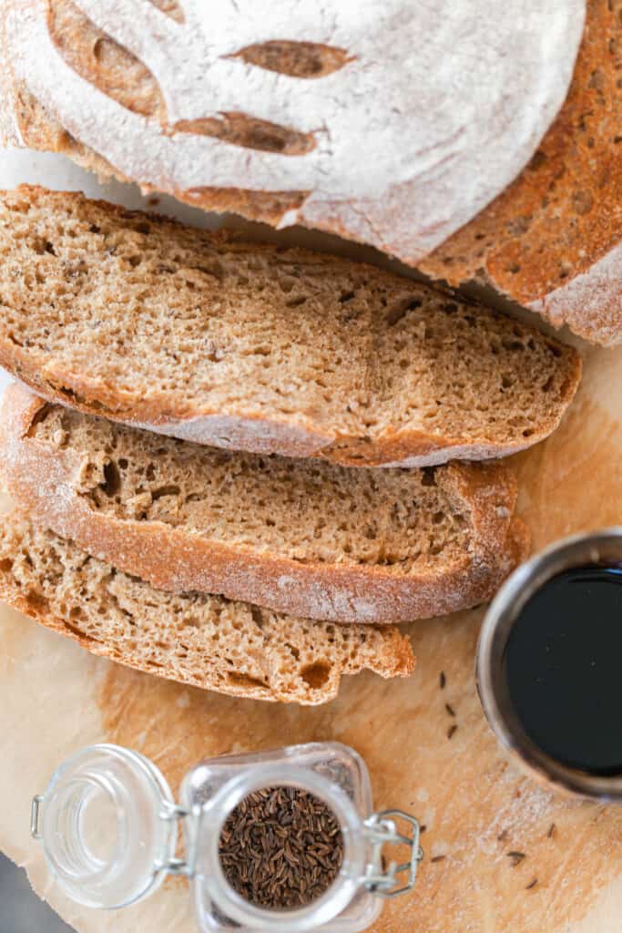 overhead photo of a loaf of sourdough rye bread sliced with the slices gently overlapping on parchment paper with a small jar of caraway seeds and a mason jar of molasses to the right