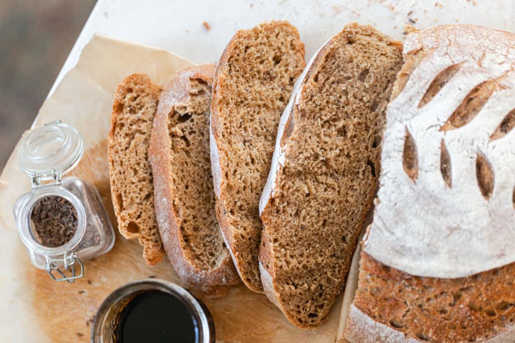 loaf of sourdough rye bread sliced on a wood cutting board with spices and molasses in little glass jars in front of the loaf