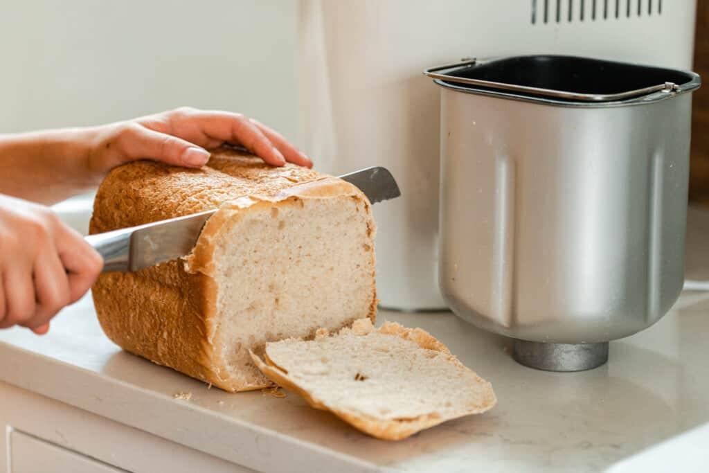 hands slicing a loaf of sourdough sandwich bread on a white quartz countertop with a bread machine pan to the right