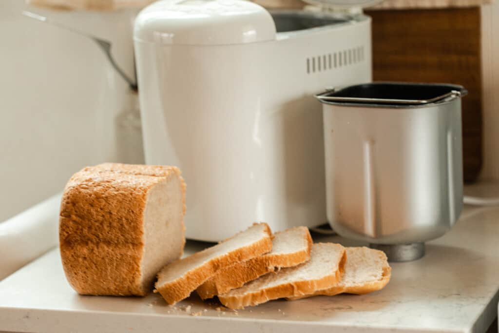 loaf of sourdough bread on a quartz countertop with half the loaf sliced. A bread machine and bread machine baking pan sit right behind the sliced loaf