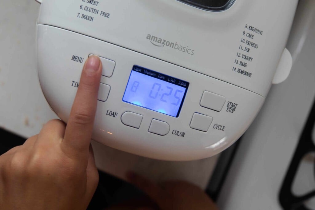 finger pushing the menu button on a bread machine to reveal kneading setting at 25 minutes.