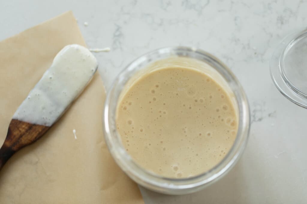 active sourdough starter in a glass mason jar on a white countertop with parchment paper and a small wooden spatula to the left.