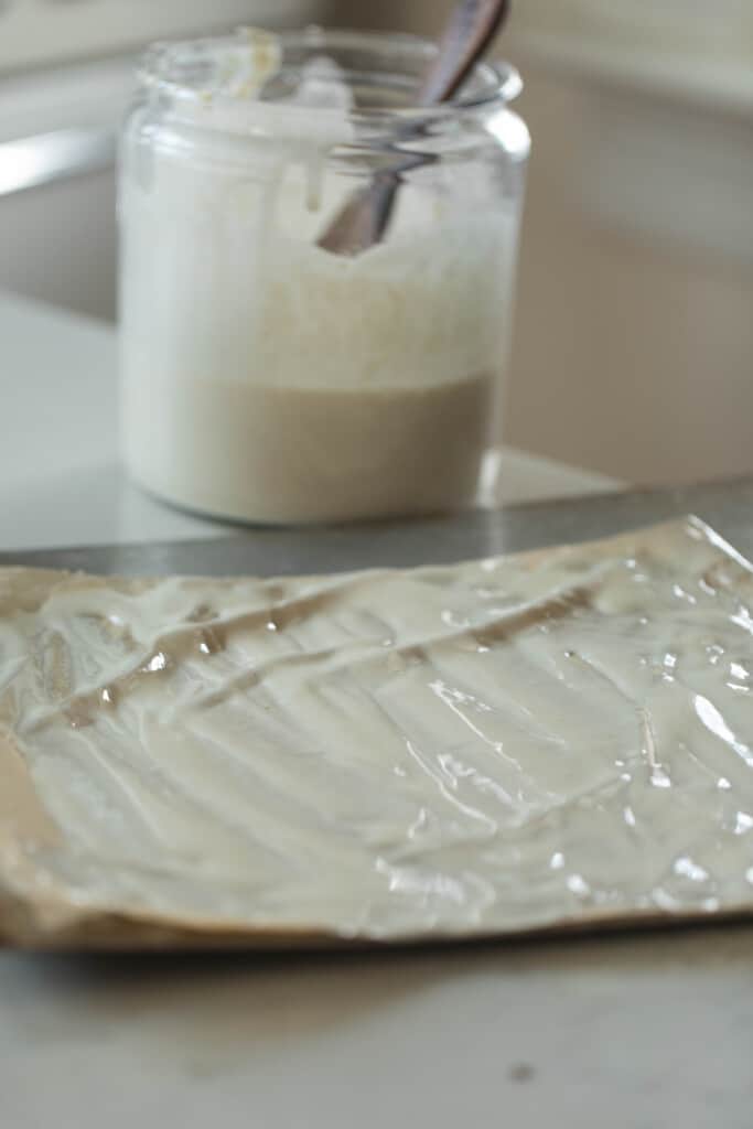 sourdough starter spread onto a parchment lined baking sheet with a large jar of starter in the background