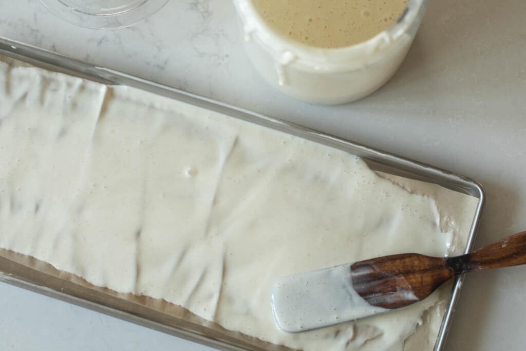 wooden spatula spreading active sourdough starer on a parchment lined baking sheet on a white quartz countertop