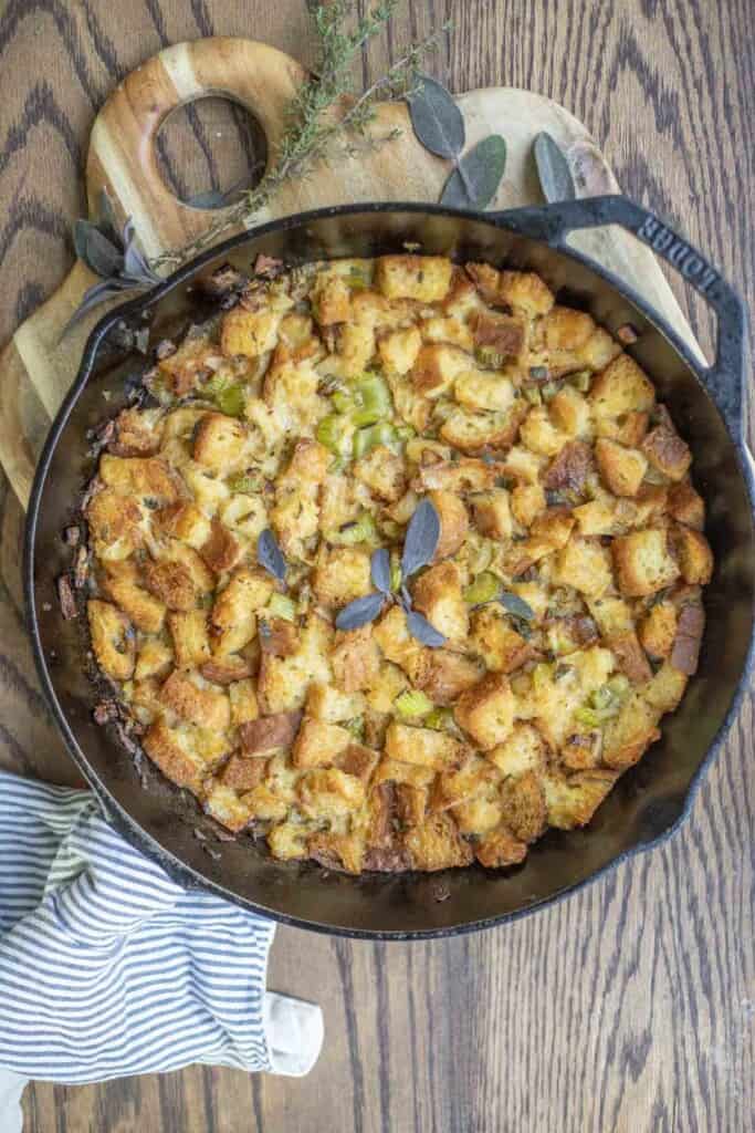 overhead photo of gluten free sourdough stuffing in a cast iron skillet on a wood cutting board with fresh herbs around the skillet