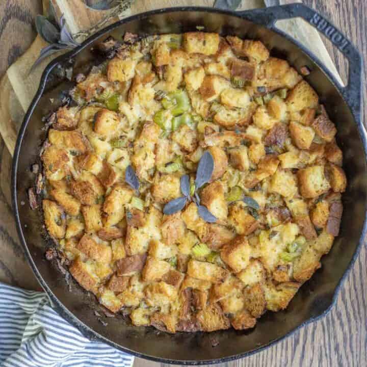 overhead photo of gluten free sourdough stuffing in a cast iron skillet on a wood cutting board with fresh herbs around the skillet