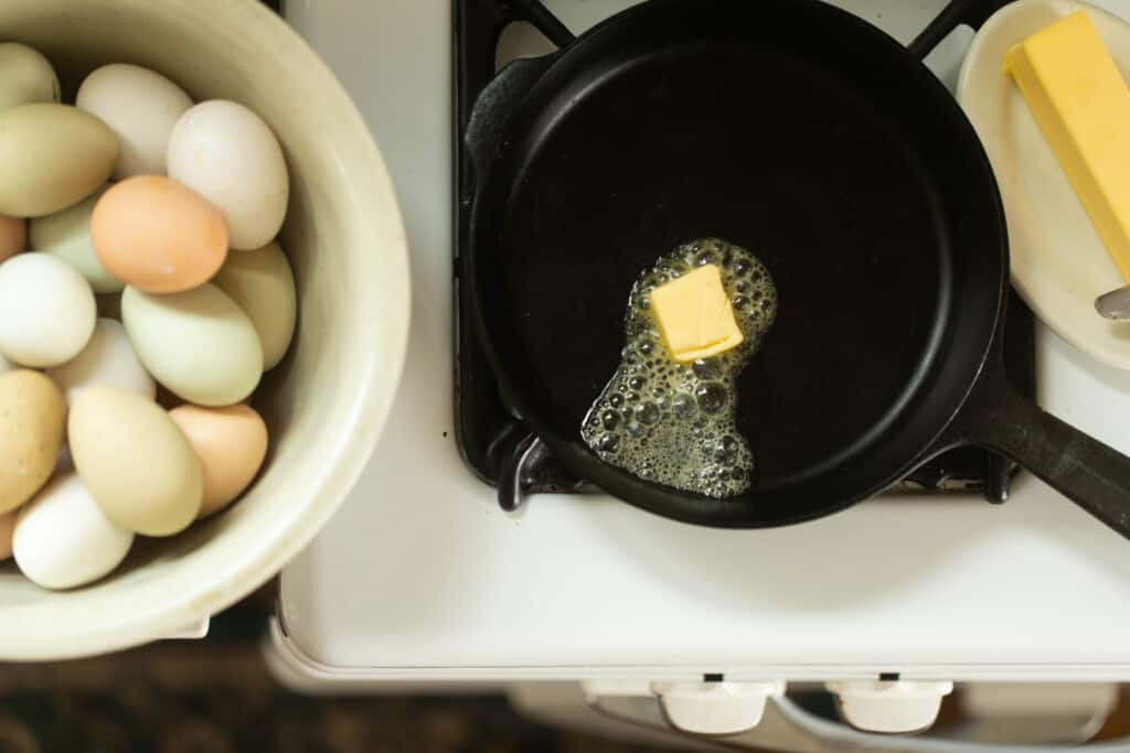 butter melting in a small cast iron skillet on a stove with a bowl of eggs to the left