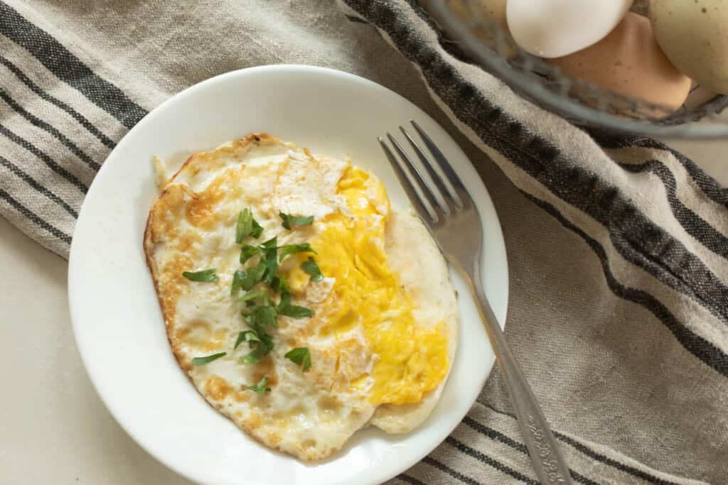 overhead photo of over hard eggs topped with fresh chives on a white plate with a fork resting on the plate. The plate sits on a white and blacked stripped towel with eggs in the back corner