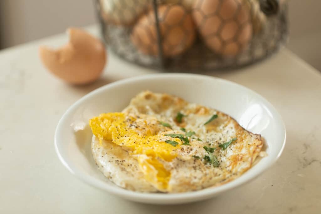 fried egg on a white dish on a white countertop with a basket of eggs in the background