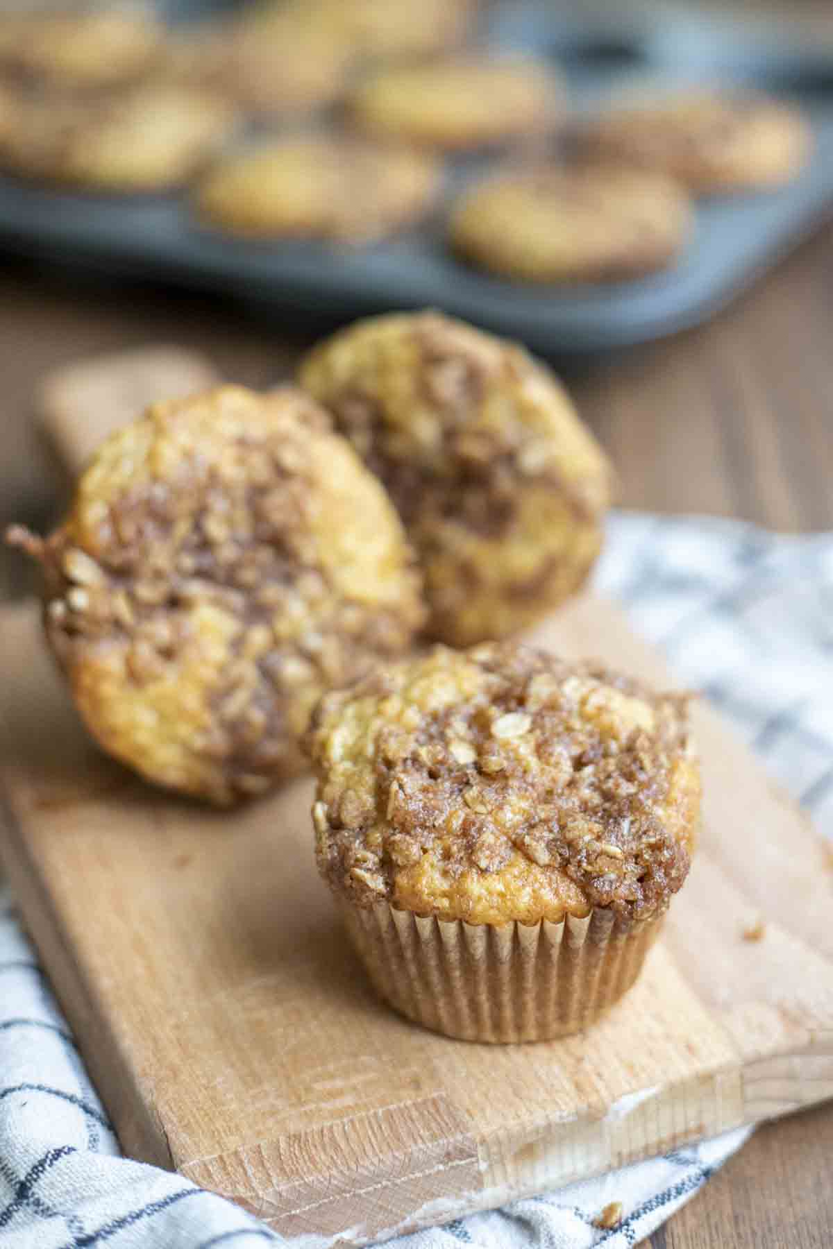 three sourdough banana muffin with a brown sugar oat crumble on a small wood cutting board with a tray of muffins in the background