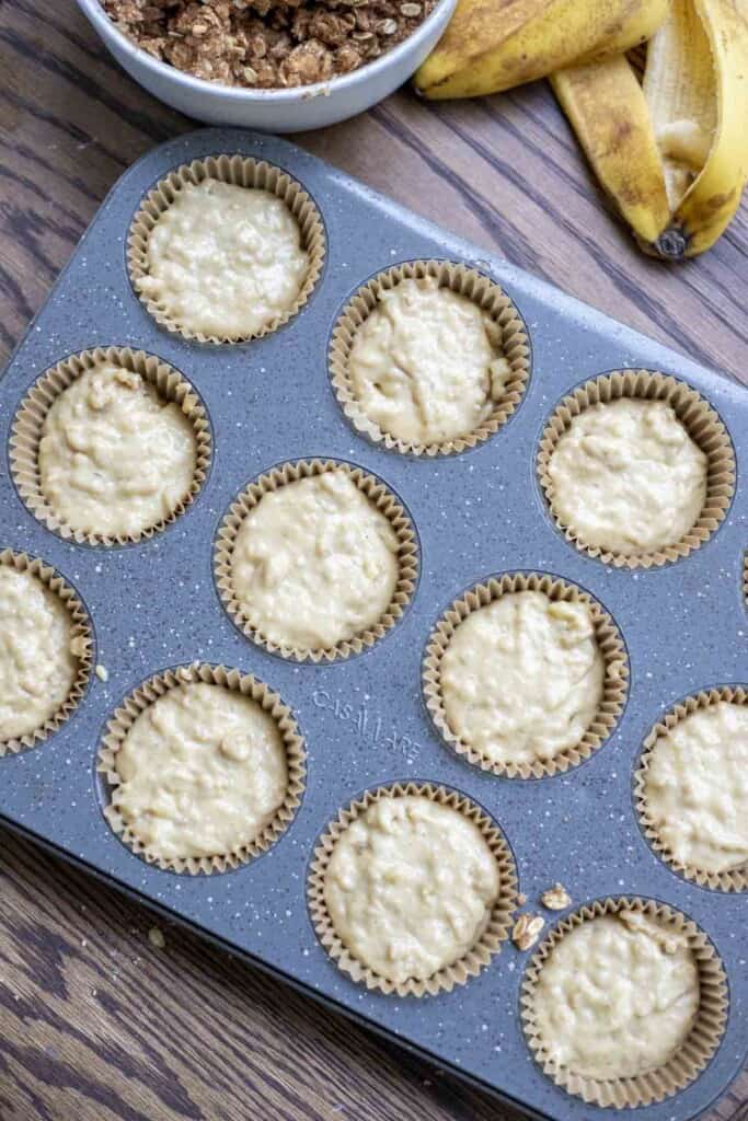 banana muffin batter in lined muffin tin with a small bowl of oat crumble and banana peels in the back corner