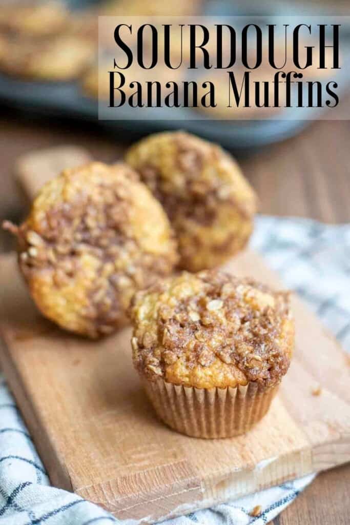 three sourdough banana muffin with a brown sugar oat crumble on a small wood cutting board with a tray of muffins in the background