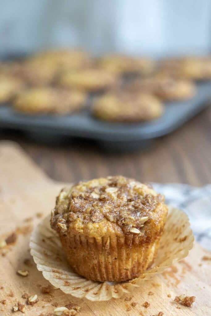 unwrapped sourdough banana muffin on a wood cutting board with crumbs surrounding the muffin. A tray of muffins are in the background