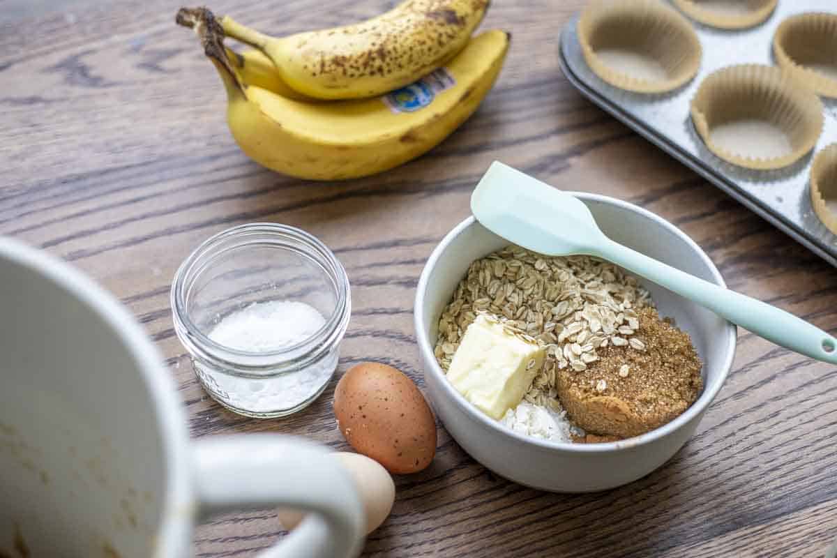 bowl of crumble ingredients, a small jar of baking powder and baking soda, ripe bananas, eggs, and a muffin pan with liners on a wood table