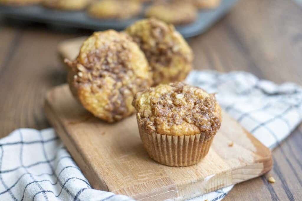 three sourdough banana muffins on a small wooden cutting board on a white an black checked towel on a wood table with a muffin pan of muffins in the background