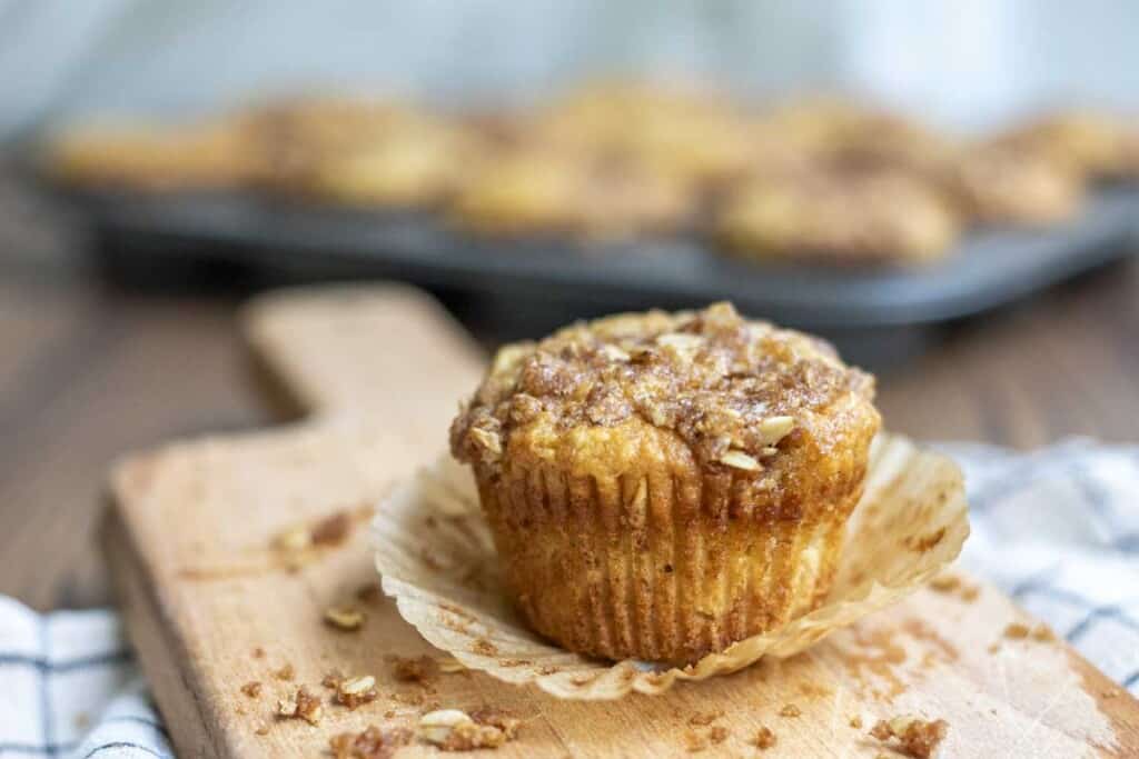 a unwrapped sourdough banana muffin with a oat crumble on a wood cutting board with oat crumble surrounding the muffin. There is a tray of muffins in the background