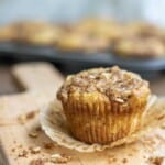 a unwrapped sourdough banana muffin with a oat crumble on a wood cutting board with oat crumble surrounding the muffin. There is a tray of muffins in the background