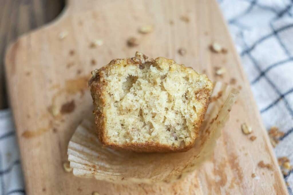 half of a sourdough banana muffin on a brown muffin liner on a wood cutting board with oat crumble sprinkled about. The cutting board is on white and black checked towel