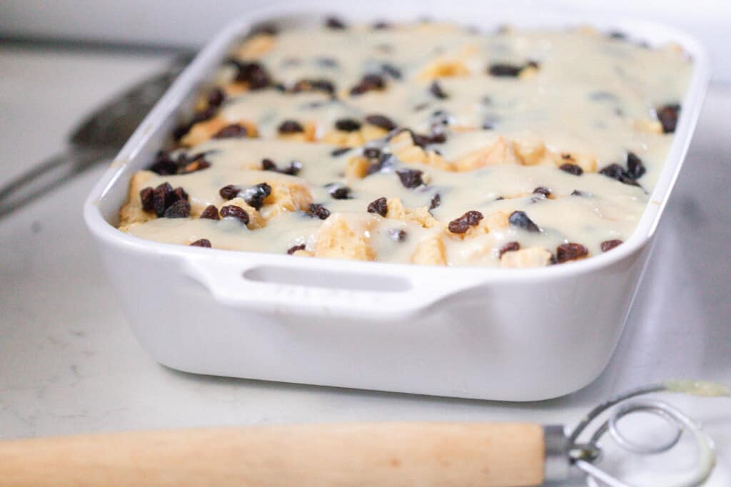 white baking dish filled with sourdough bread pudding topped with raisins and a bead pudding sauce. The baking dish sits on a white countertop with a wooden dough whisk in front on the dish