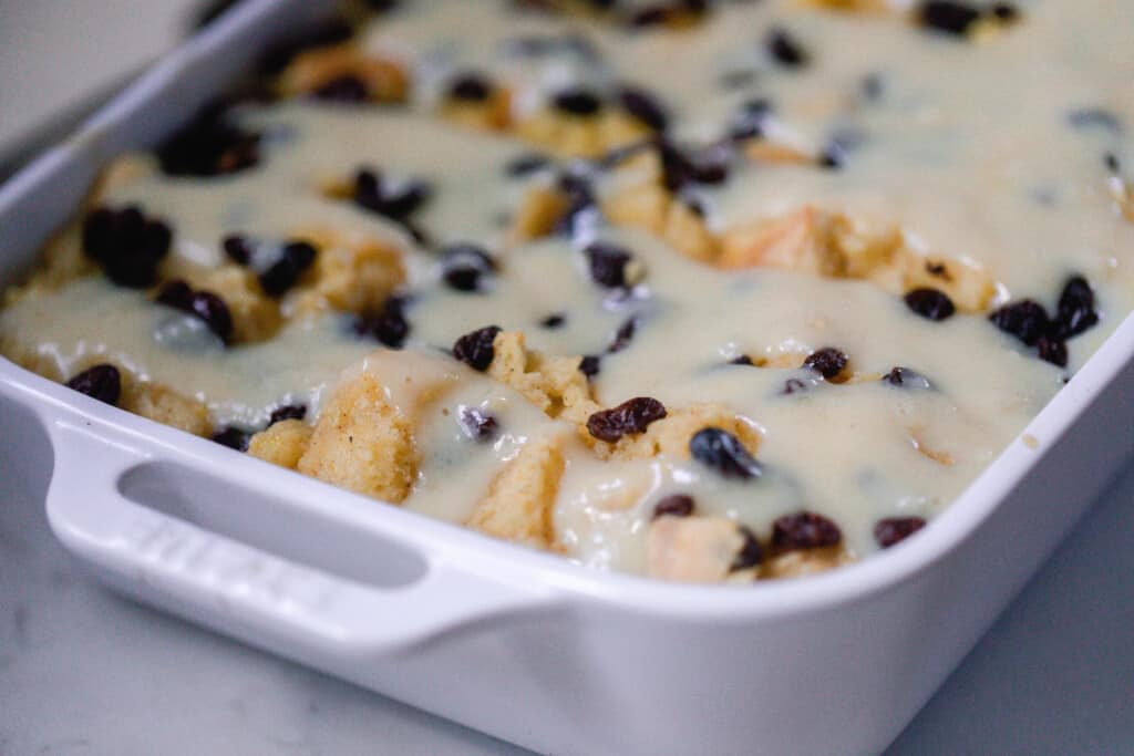 side corner of a white baking dish full of bread and butter pudding topped with raisins and a creamy sauce