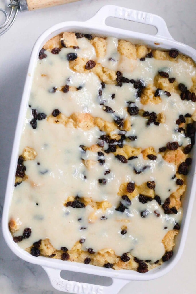 overhead photo of a white baking dish full of sourdough bread pudding topped with raisins and a bread pudding sauce. The dish sits on a white countertop 