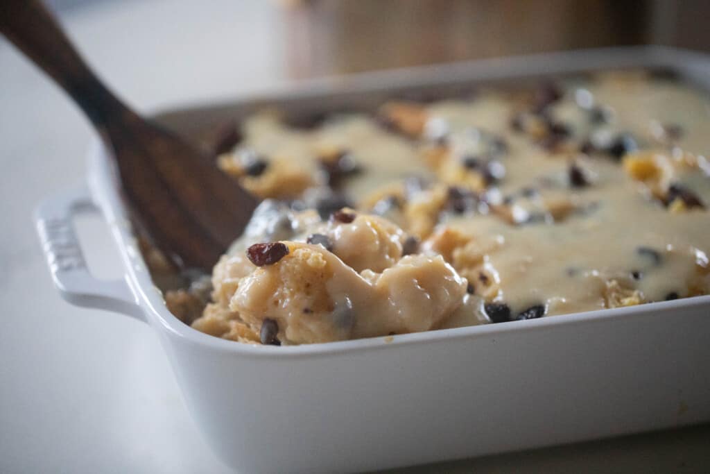 wooden spatula scooping out brioche bread pudding with a creamy sauce and raisins in a white baking dish