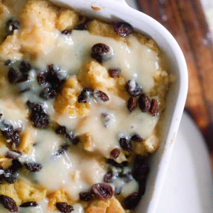 overhead photo of sourdough bread pudding topped with raisins and a creamy sauce in a white baking dish on a white countertop with a wooden spatula to the right
