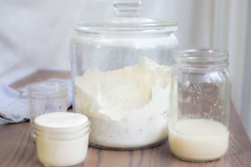 glass jars full of flour, sourdough starter, a water yeast mixture and salt on a wood table