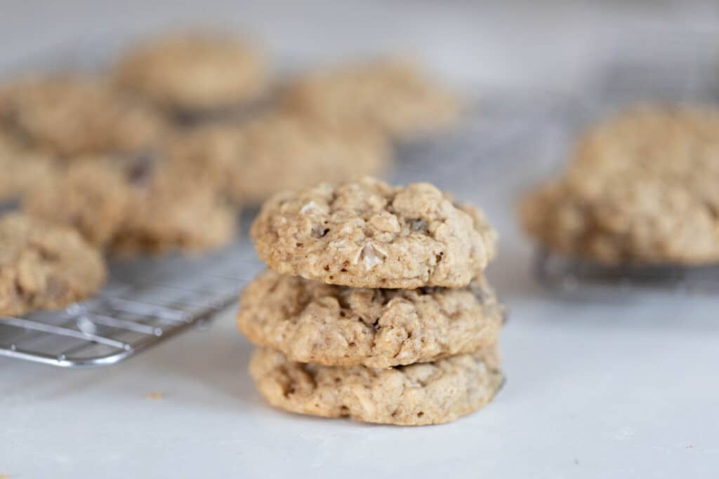 three sourdough oatmeal cookies stacked on top of each other on a white quartz countertop with more cookies on wire racks in the background
