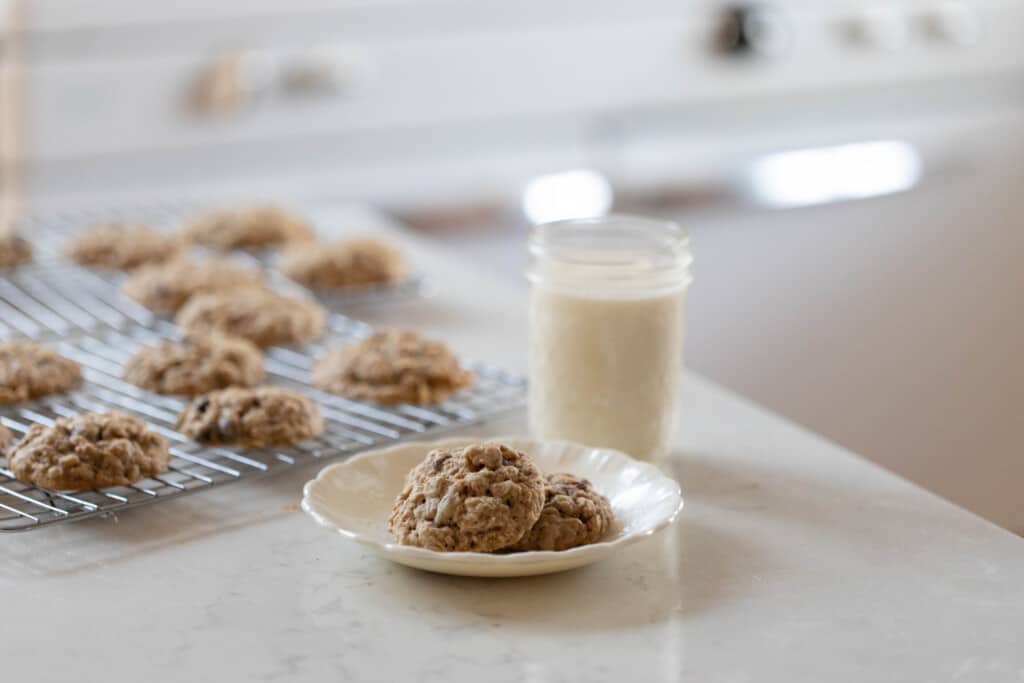 two sourdough oatmeal raisin cookies on a white plate with a class of milk and a wire rack of more cookies on a white quartz island