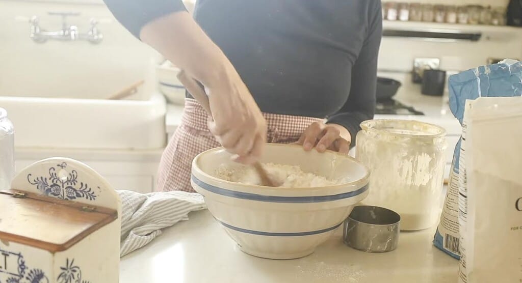 woman wearing a black long sleeve shirt mixing a bowl of flour with a wooden spoon. Ingredients are surrounding the bowl