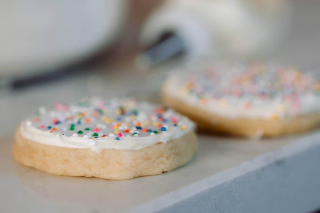 two round sourdough sugar cookies with white icing topped with rainbow sprinkles. The cookies sit on a white countertop with an icing bag in the background