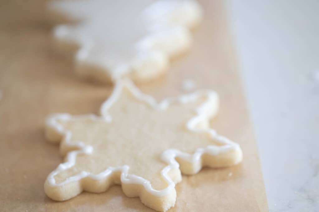 close up of a snowflake shaped sourdough sugar cookie with icing around the edges with another tree sugar cookie with white icing in the background. Both cookies lay on parchment paper