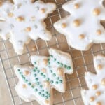 sourdough sugar cookies cut out as snowflakes and trees decorated with white frosting and gold balls. on a wire rack on parchment paper