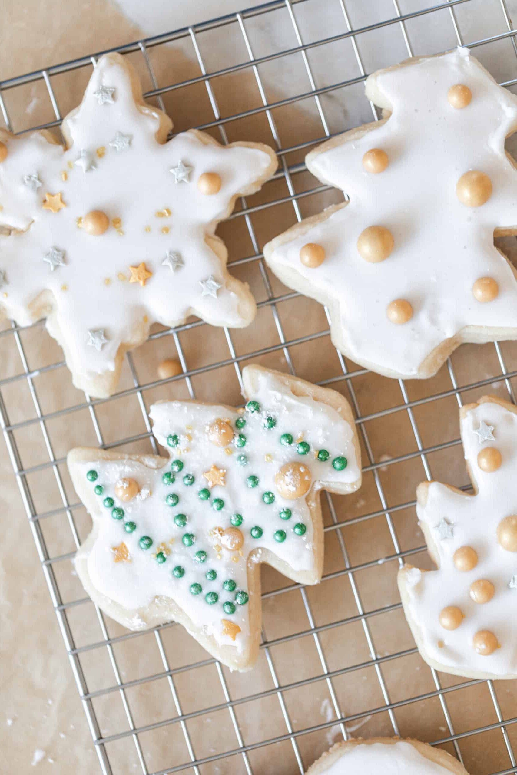 sourdough sugar cookies cut out as snowflakes and trees decorated with white frosting and gold balls. on a wire rack on parchment paper