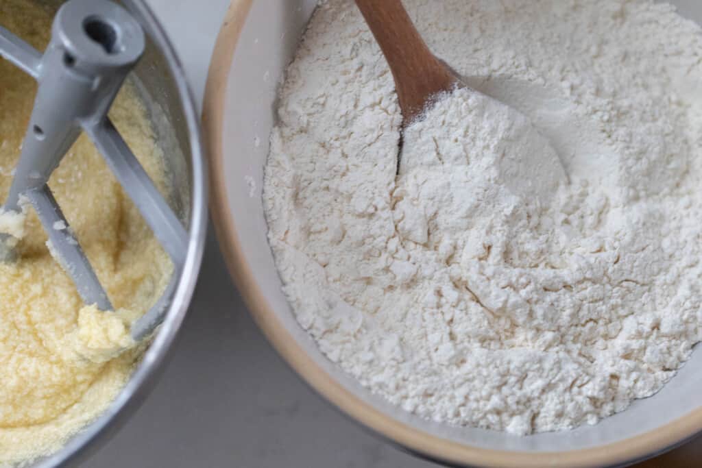 overhead photo of flour in a boll with a wooden spoon next to a stand mixer bowl of whipped butter and sugar with the paddle attachment in the bowl