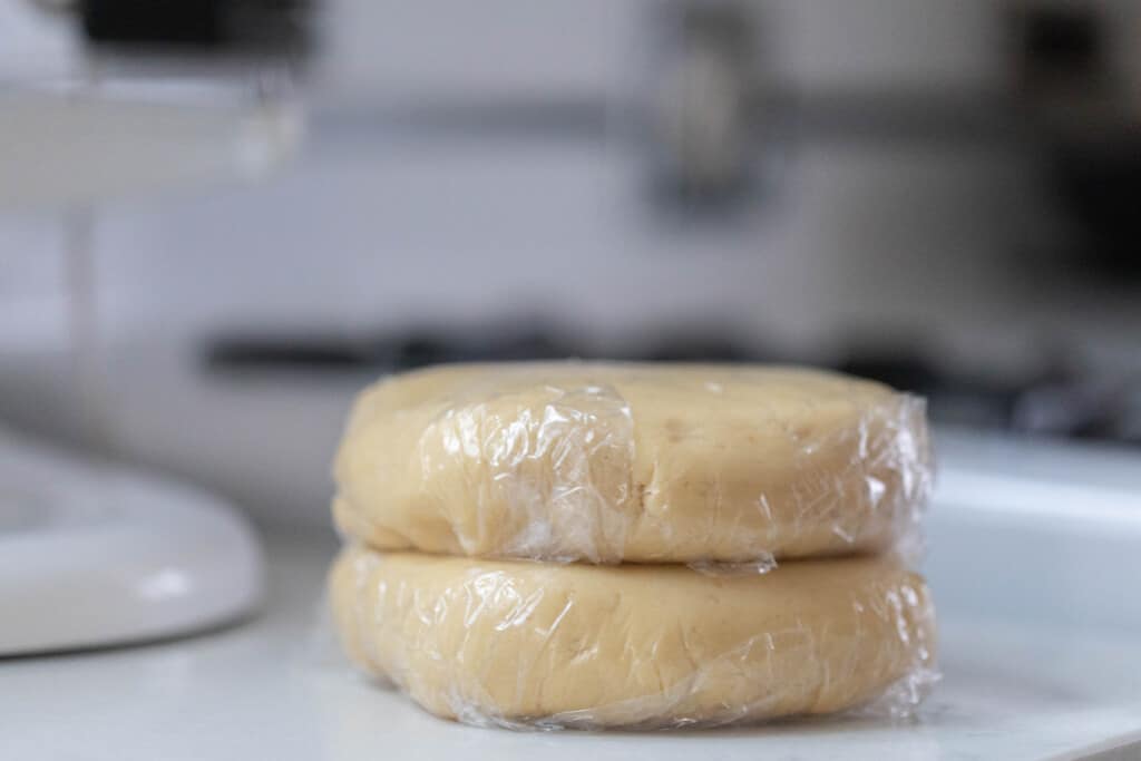 two disks of sourdough sugar cookie dough wrapped in plastic wrap on a white countertop