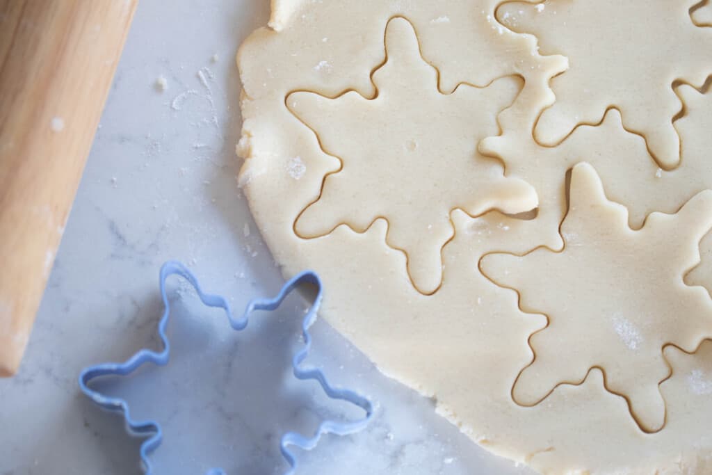 sourdough sugar cookie dough rolled out on a quartz countertop with snowflake cookie cutter shapes cut out in the dough. The cookie cutter and a rolling pin are to the left of dough