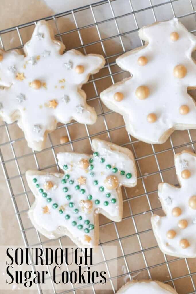 sourdough sugar cookies cut out as snowflakes and trees decorated with white frosting and gold balls. on a wire rack on parchment paper