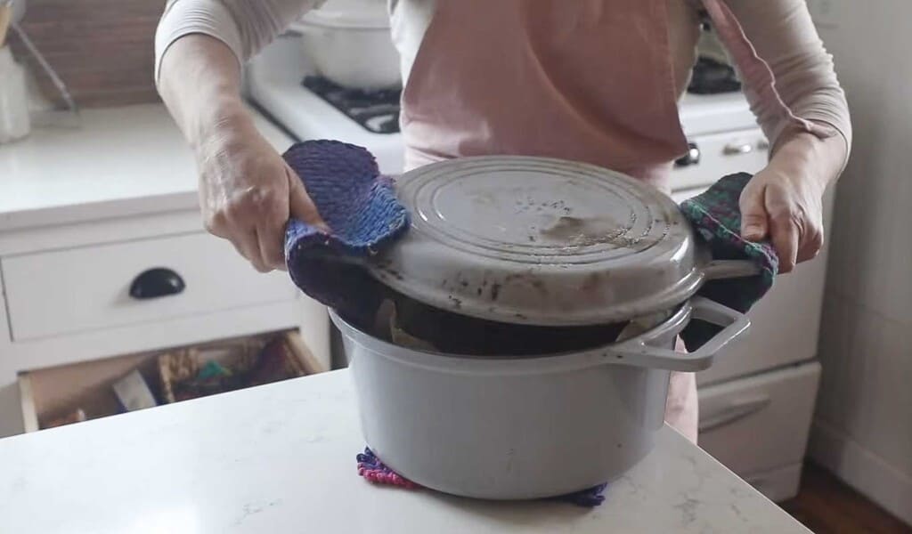 a woman wearing a pink apron adding a lid to a dutch oven that is sitting on a white quartz countertop