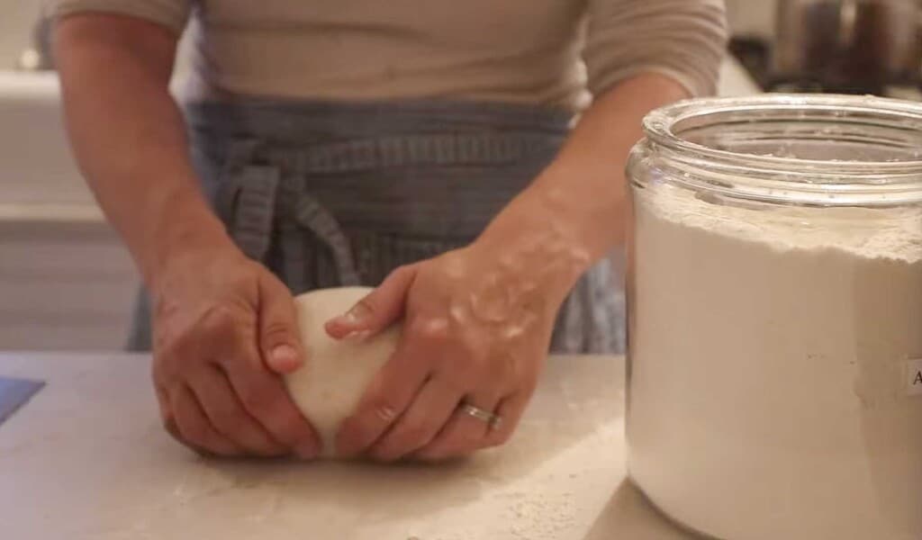 a woman wearing a blue apron pulling a boule of spelt sourdough bread dough towards her against the white countertop to create tension in the dough. A large jar of flour is to the right.