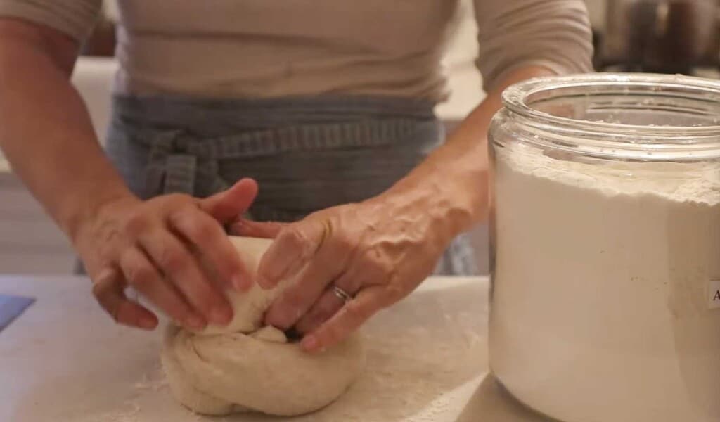 hands rolling up a loaf of sourdough spelt bread on a white countertop with a large jar of flour to the right