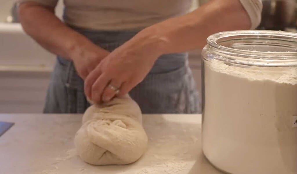 a woman in a blue apron creating a shoelace patter on bread dough on a white quartz counter with a large jar of flour to the right