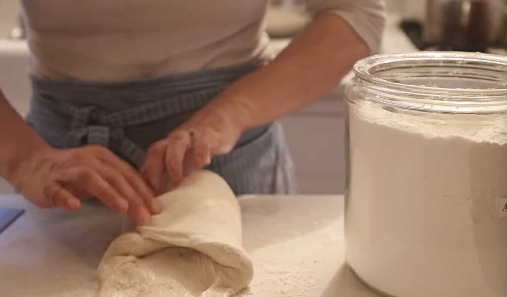 a woman wearing a blue and white apron folding sourdough spelt bread on a white countertop with a large jar of flour to the right