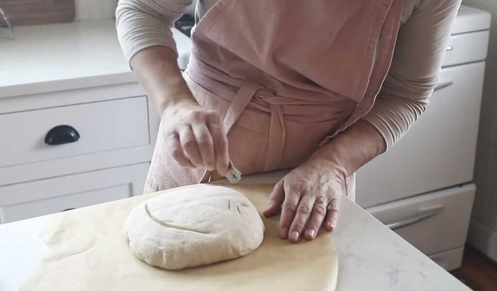 a woman wearing a pink apron scoring a loaf of spelt sourdough bread on parchment paper on a white countertop