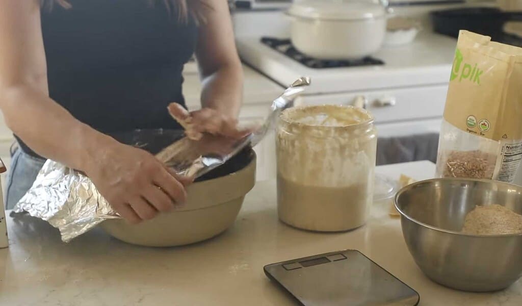 a woman placing a piece of tin foil on top of a stoneware bowl with a jar of sourdough starter to the right and a bowl of flour