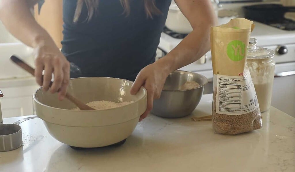 a woman wearing a black top stirring bread dough together with a wooden spoon in a stoneware bowl on a white countertop.