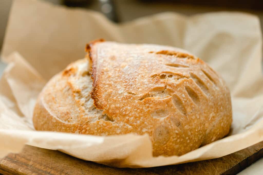 close up of a boule of spelt sourdough bread with a wheat pattern baked into the loaf. The loaf of bread rests on parchment paper on a wooden cutting board.