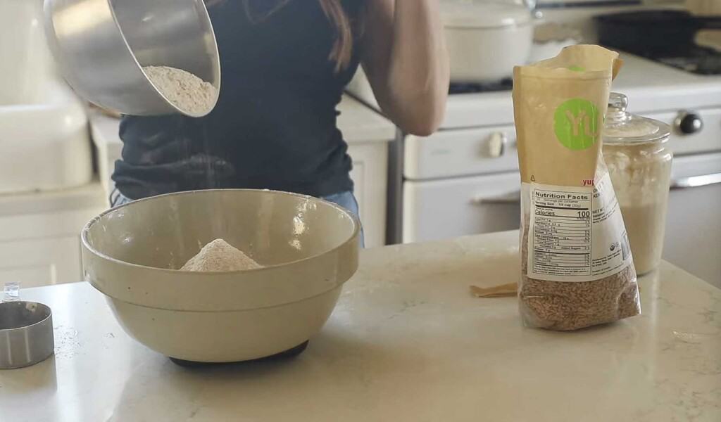 woman wearing a black shirt pouring flour from a bowl to another bowl on a white countertop